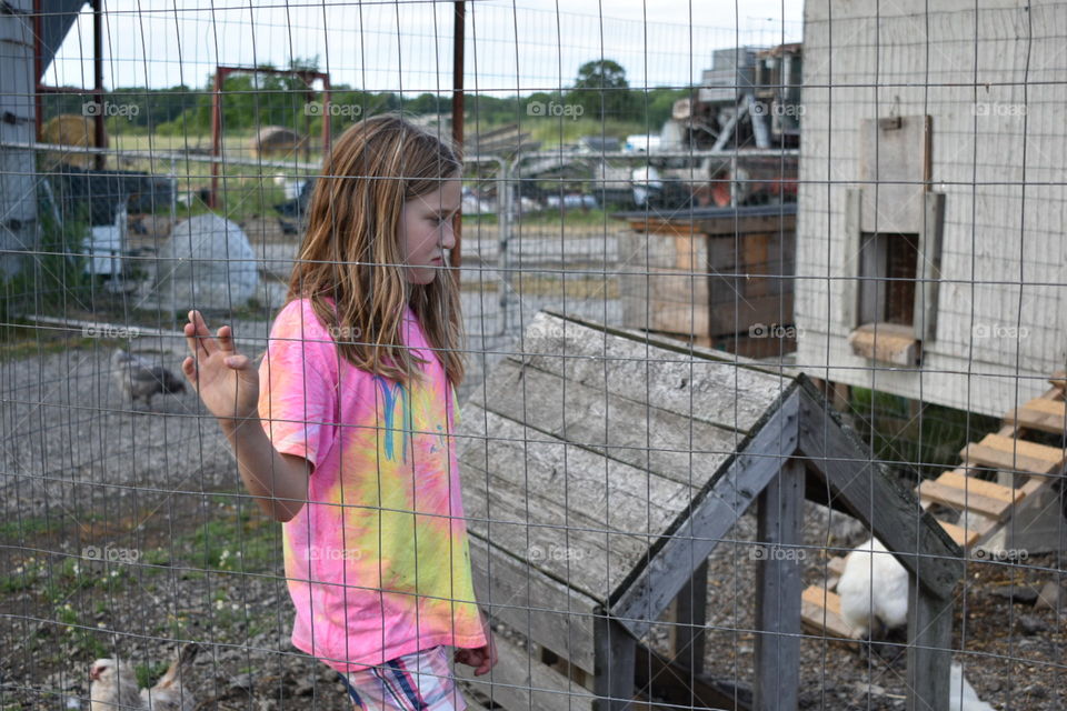 A child has a melancholy look on her face as she holds on the the fencing around her chicken coop on her grandparents farm. She stands it in a bright pink and yellow tie-die shirt agains the neutral background colours. 
