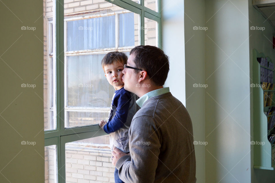 Father kissing toddler son on the cheek in front of a bright window indoors