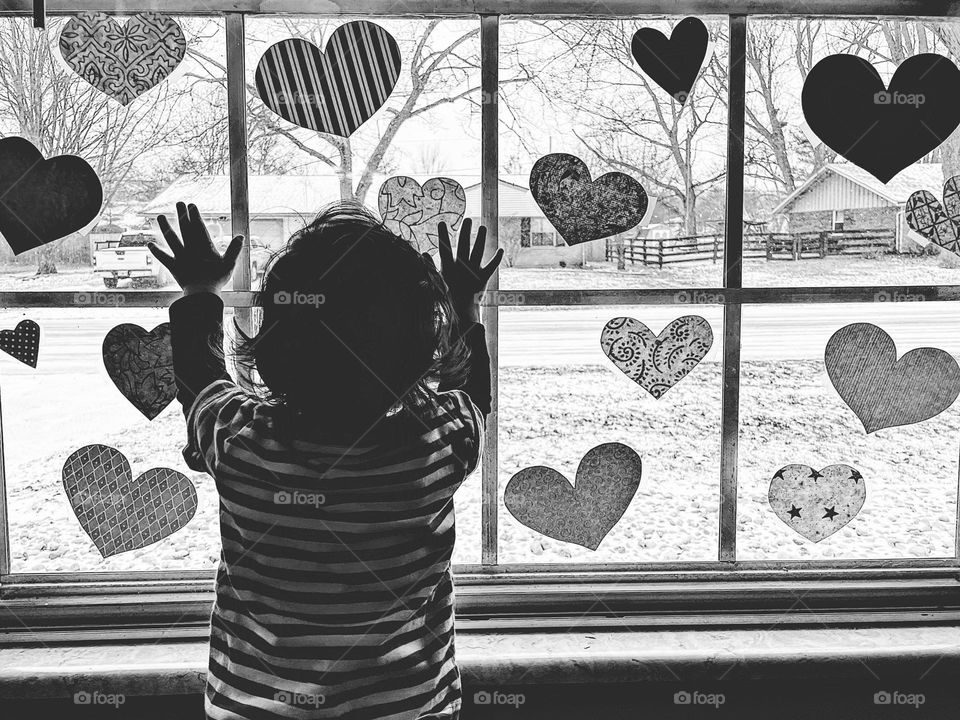Baby at window with hands reaching out for heart decor, baby looks out window with Valentine’s Day decorations on it,  watching the snow, baby looks out of window, black and white portrait, monochrome image of baby in window, people in windows 