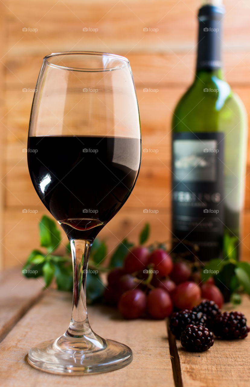 glass of red wine on wood table next to red grapes and blackberries with the wine bottle blurred in the background with a wood wall