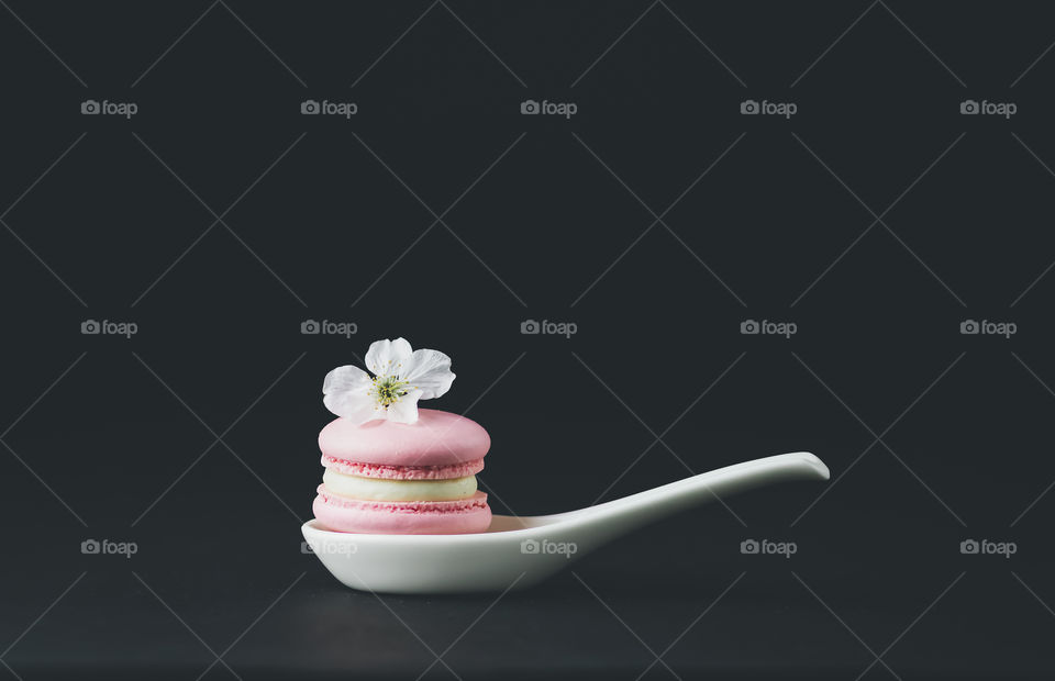 Selective focus to one pink macarons with vanilla cream and  white flower on the spoon at dark background. Copy space.