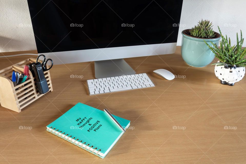 Desk with stationary, computer, pot plant and “Happy Thoughts & Positive Plans” journal