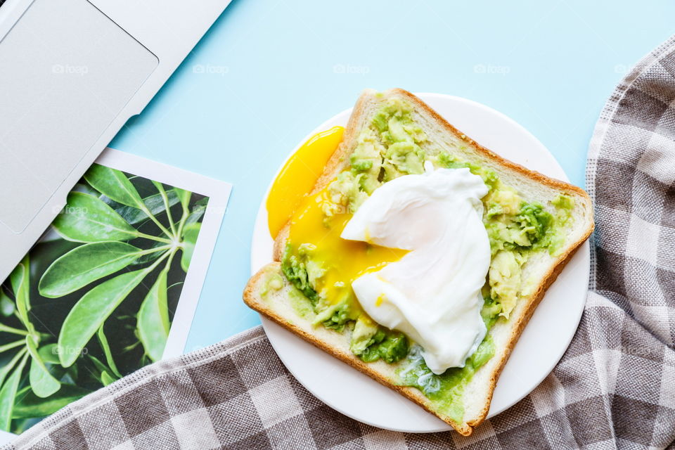 Sandwich with fresh green avocado, poached egg on white plate lying on blue background 