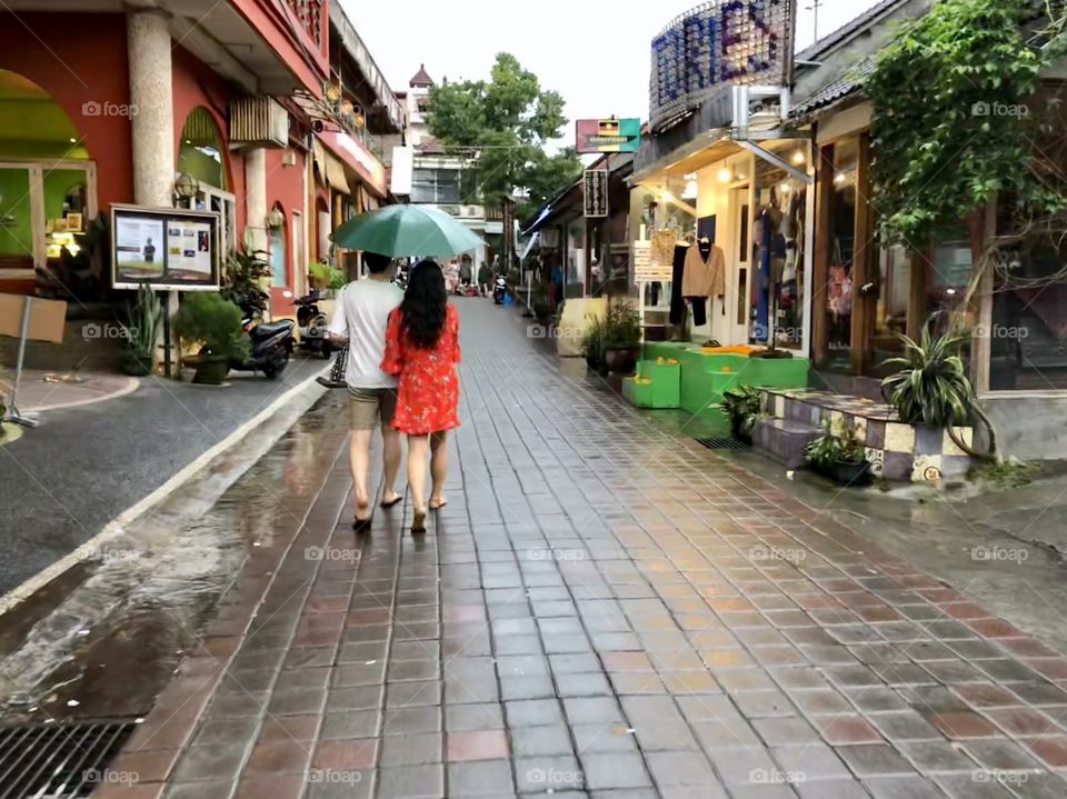 Walking in stride 1. Adult couple with open umbrella. Brick paved street. Shops. Ubud, BalI. 2018.