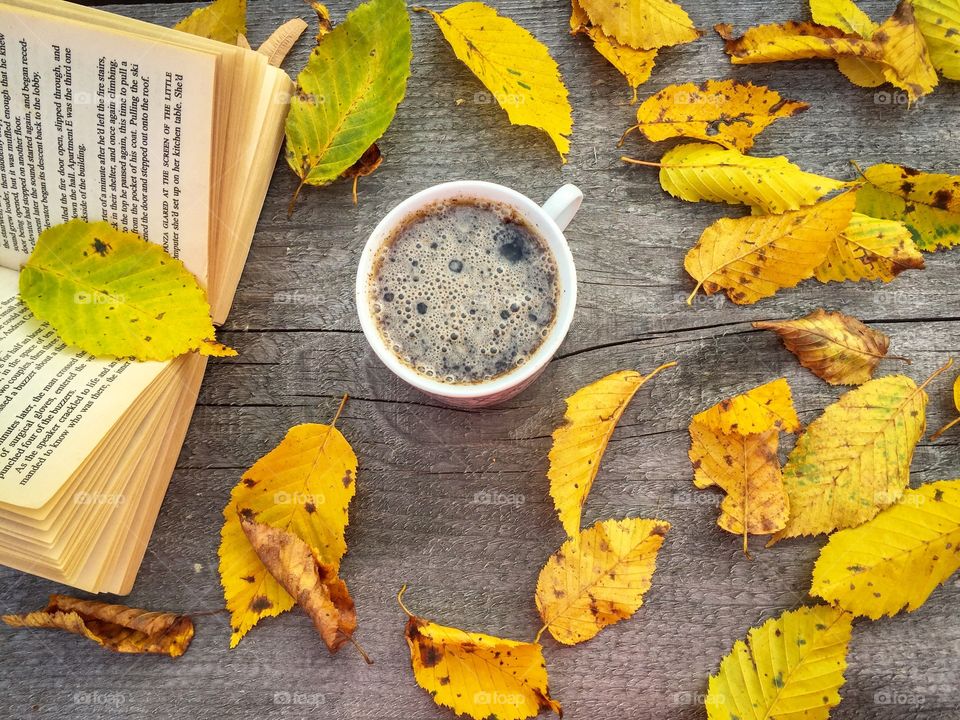 Cup of coffee on wooden table with a book and yellow autumn leaves beside