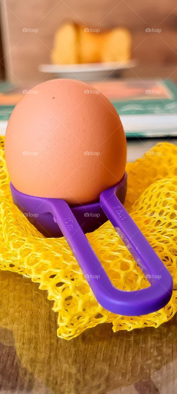Purple kitchen utensil with an egg inside, wrapped in a yellow ecological mesh bag. In the background, a blurred image of a cookbook and a homemade cake.