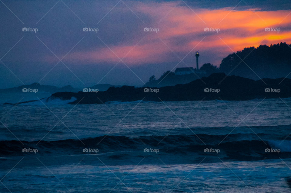 Silhouette of the Northwest Pacific Coast with waves splashing on the sandy beach & on the rocky outcrops. A lighthouse shines it’s light out to sea to warn mariners of the hazardous rocks & the sun sets in the cloudy but colourful sky. 