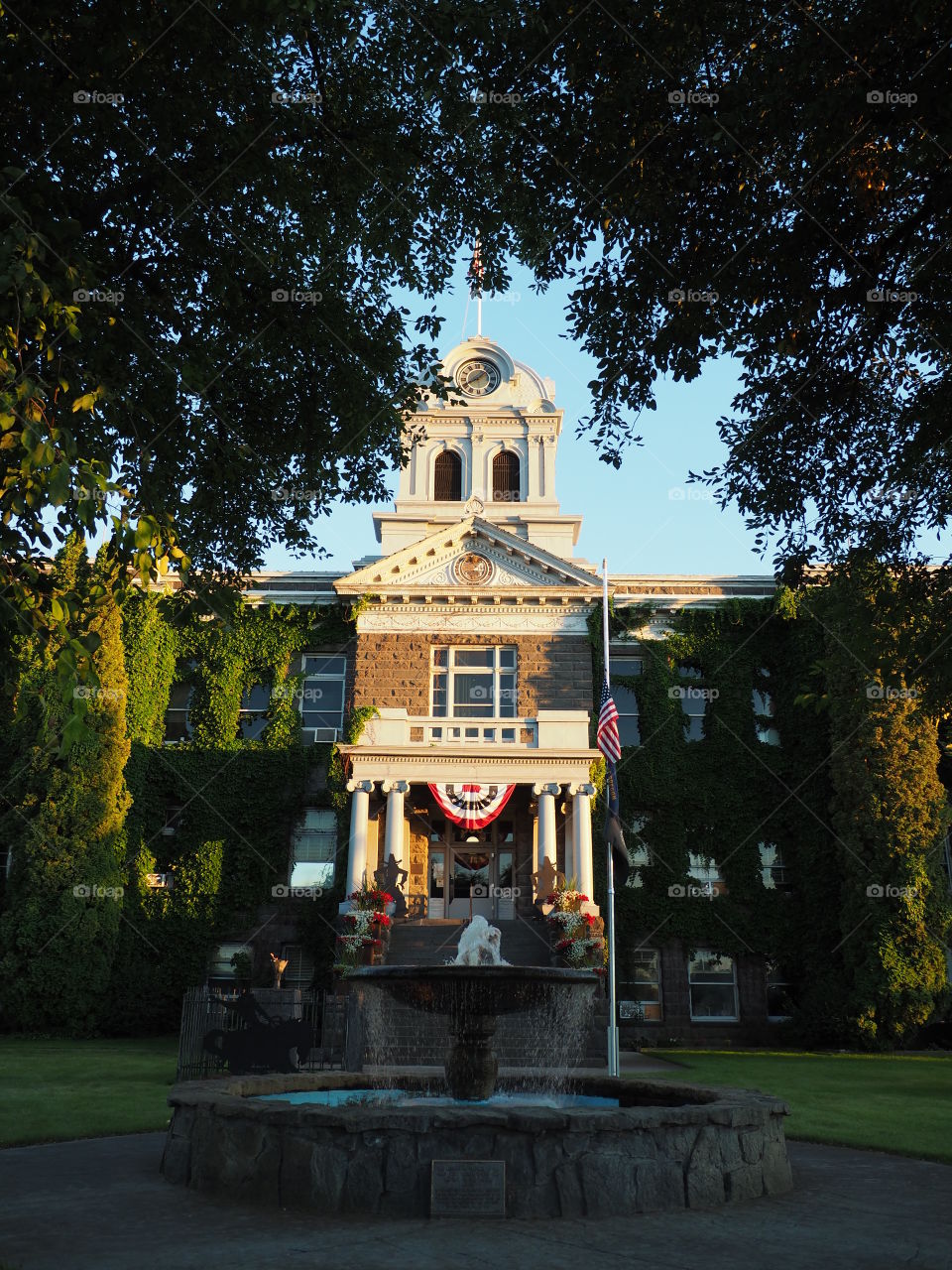 The old Crook County Courthouse in Central Oregon is beautifully lit as the sun goes down. The railings on the steps have flower baskets, the American flag flies at half- mast, and the fountain flows. 