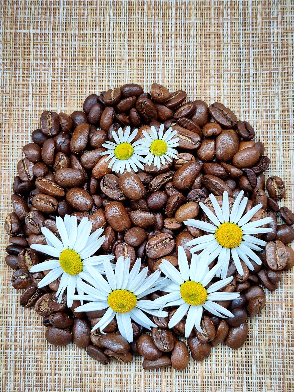 Roasted coffee beans in the form of a circle.  On the coffee circle, eyes and a smiling mouth of daisies