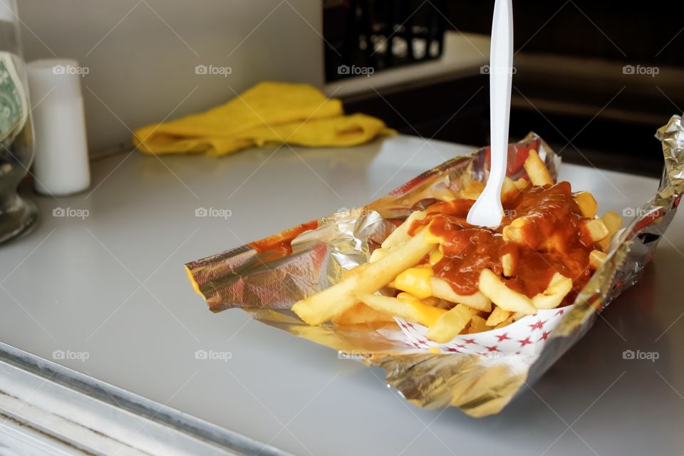 Chili cheese fries hot and ready sit on a vendor counter. The messy festival food is served with aluminum foil under and a plastic fork sticking straight up. 