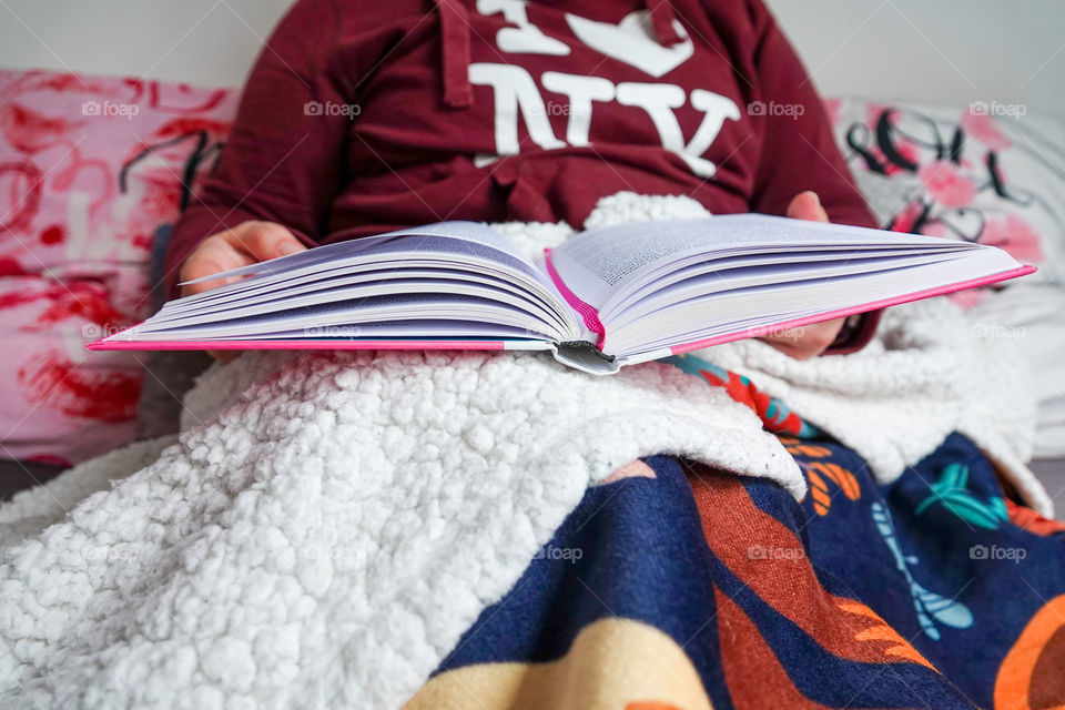 A young girl is reading a book in bed wrapped in a comfortable warm blanket.