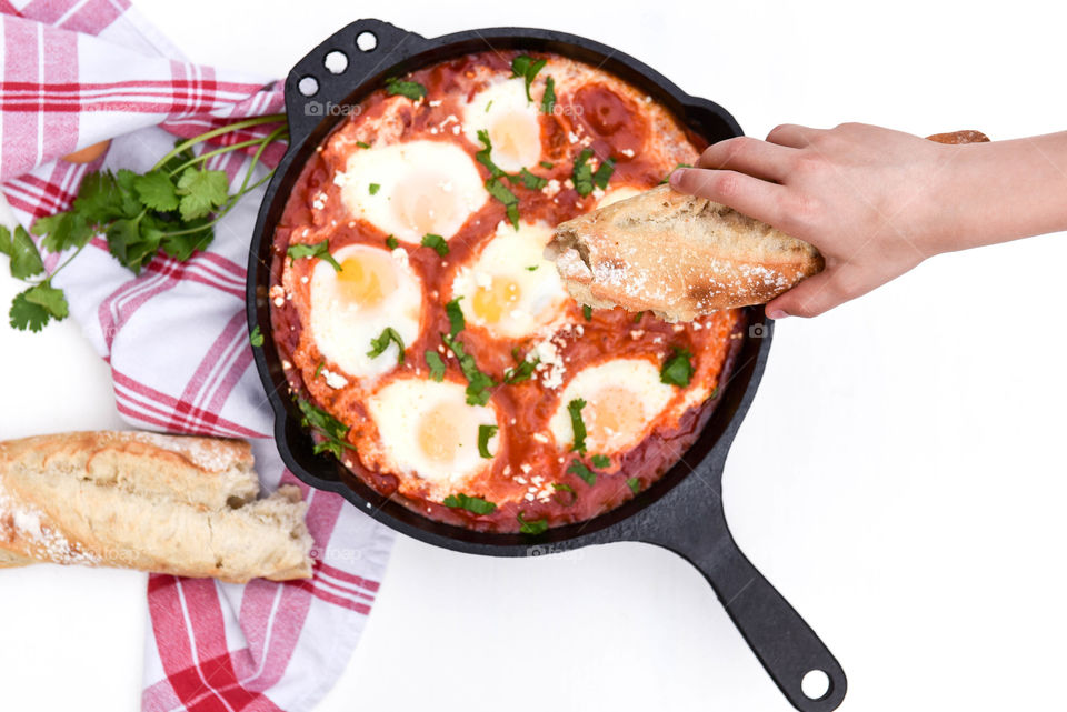 Person's hand holding a baguette over a skillet shakshuka dish