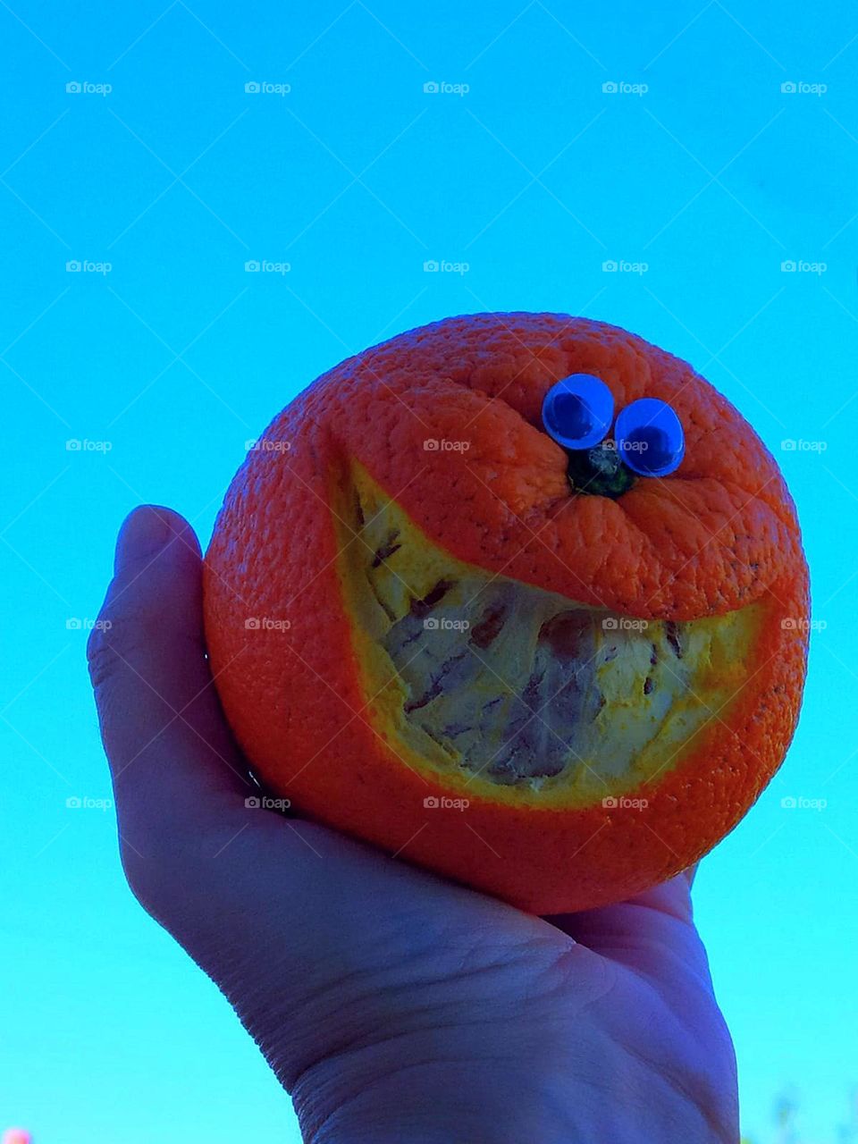 Fruit.  A woman's hand against a blue sky holds a smiling orange