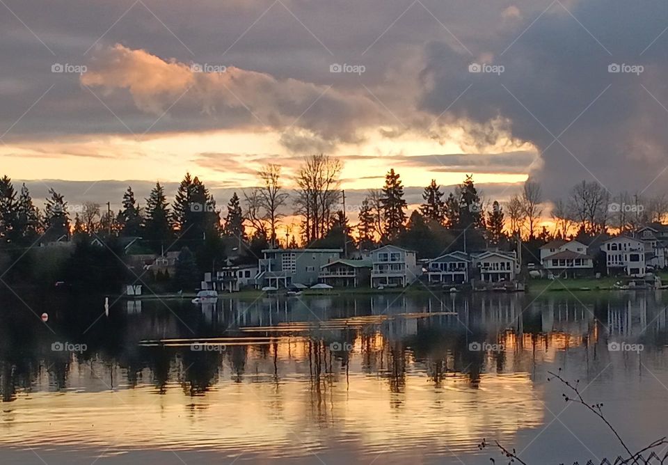 A background of luminous clouds,  and a beautiful orange and pink sunset, tree-lined with the waterfront homes mirrored on the lake.