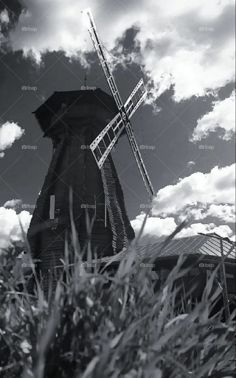 Black and white photo.  Bottom view.  In the foreground, grass through which you can see: an old wooden mill and a sky with clouds