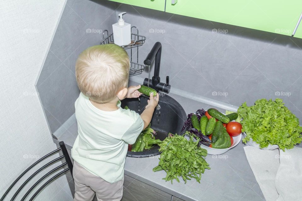 A small child washes fresh, green, vegetarian vegetables under the tap in a black sink to prepare salads and other dishes in a homemade gray and green kitchen.