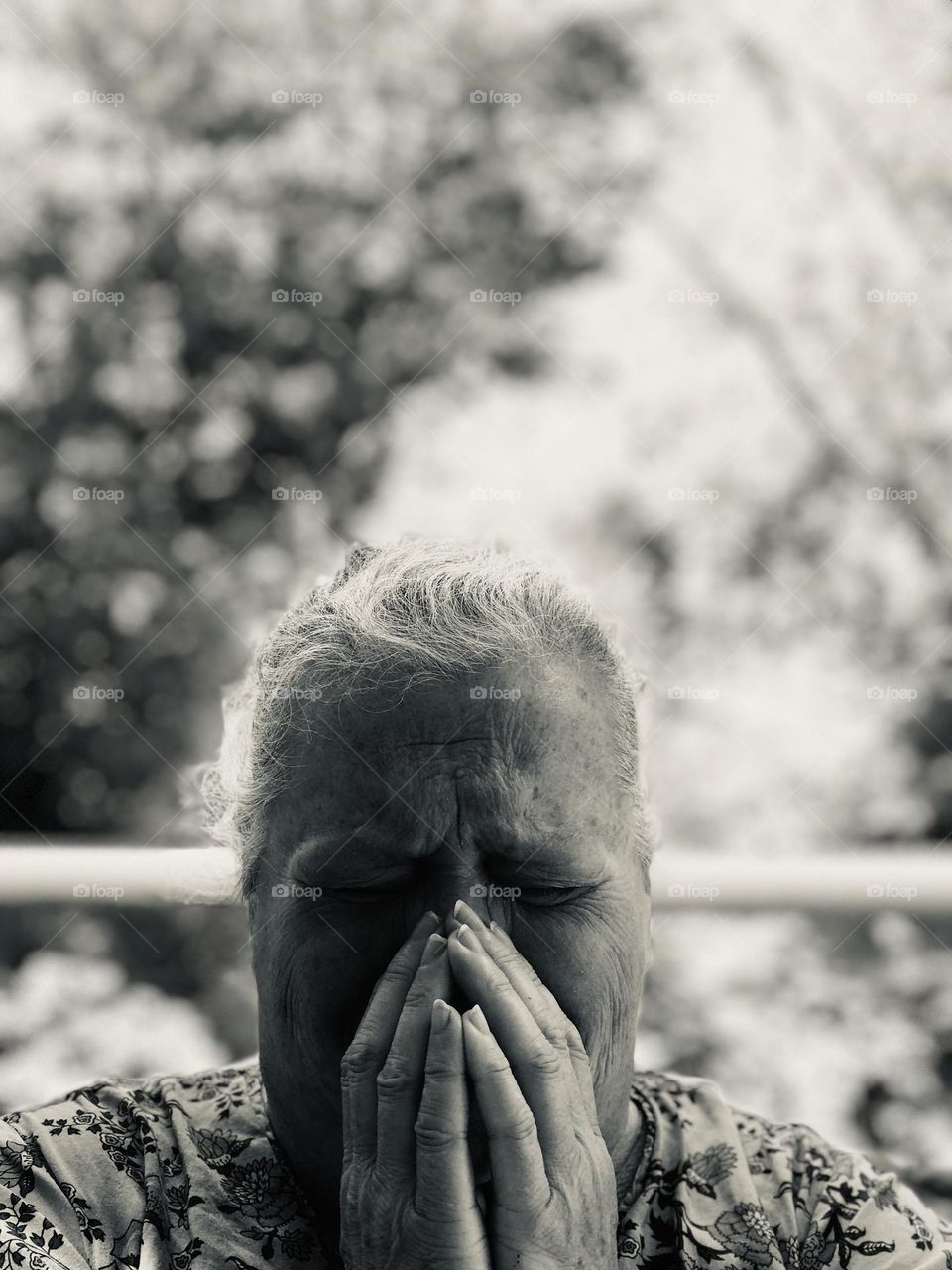 Black & white self portrait of a woman in pain. She describes the overall pain as being “so bad as if I was hit by a bus & there’s a stake going through my head”. She sits crying in pain in front of a beautiful yard, hands grasping her mouth & nose. 