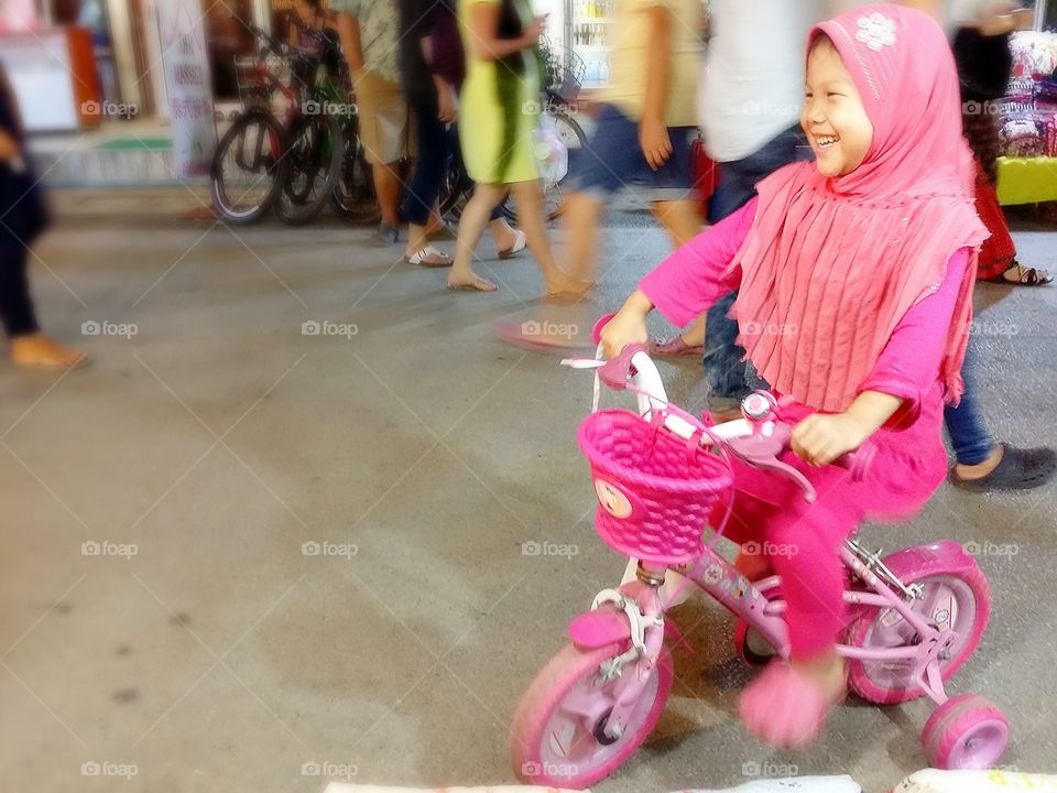 Enjoys. The kid is enjoy on pink bicycle at walking strret night market.