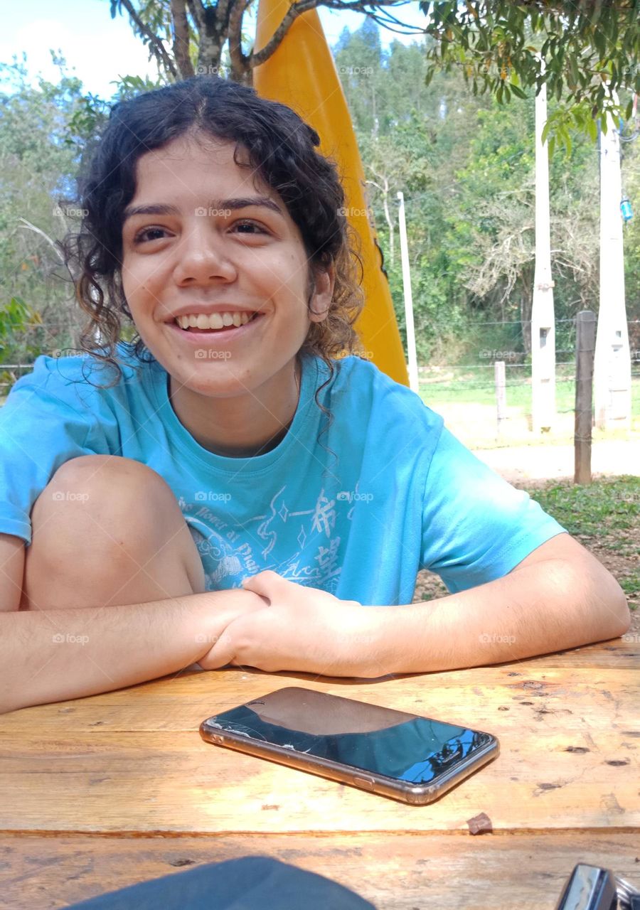 Young girl, sitting and smiling with arms over a table. Curly hair, blue t-shirt, natural background with trees, a phone on the table