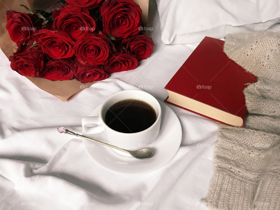 Coffee cup, red roses and red book in white bed 