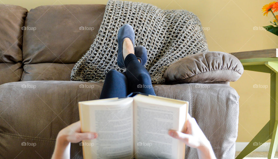 Woman laying on the floor of living room with legs propped on a couch while reading a book