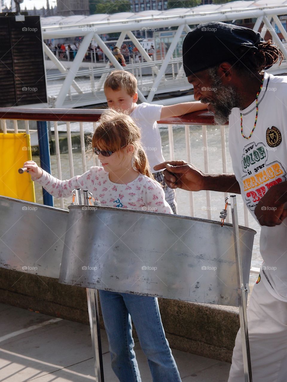 A young girl plays on the steel drums with instruction from a musician on a summer day outdoors. 
