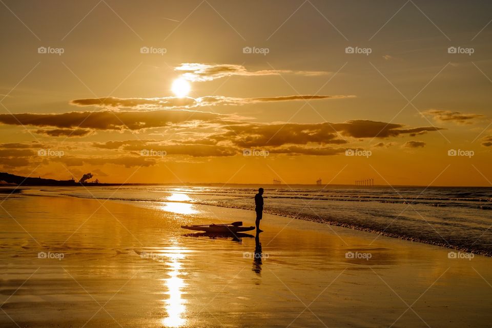 Standing on the Edge. Silhouette of a man with a sea kayak on a beach during a golden sunset that is reflected onto the wet sand. 