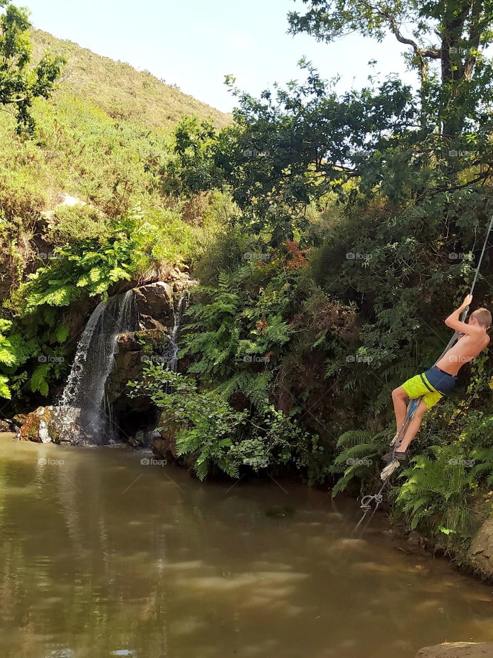 Summer and holidays are adventure! During a hike, discovery of a small waterfall. A boy is having fun swinging on a rope hanging from a tree like Tarzan.