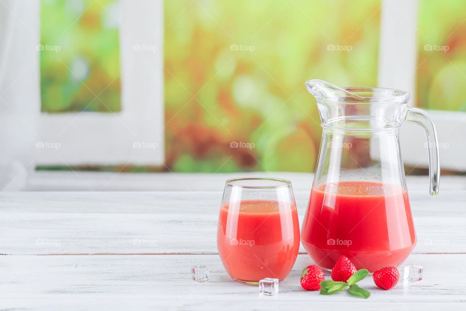 Fresh strawberry drink in a decanter and a glass with fresh berries, mint leaves and ice cubes on a white shabby table in front of a blurred window with a flowering field. Summer drinks concept.