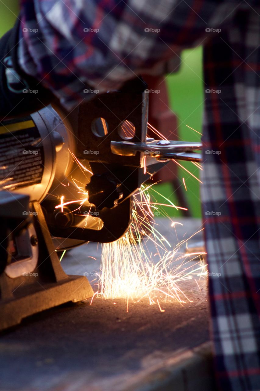 Side view of sparks from a bench grinder being used by a man on a steel part he is holding with pliers
