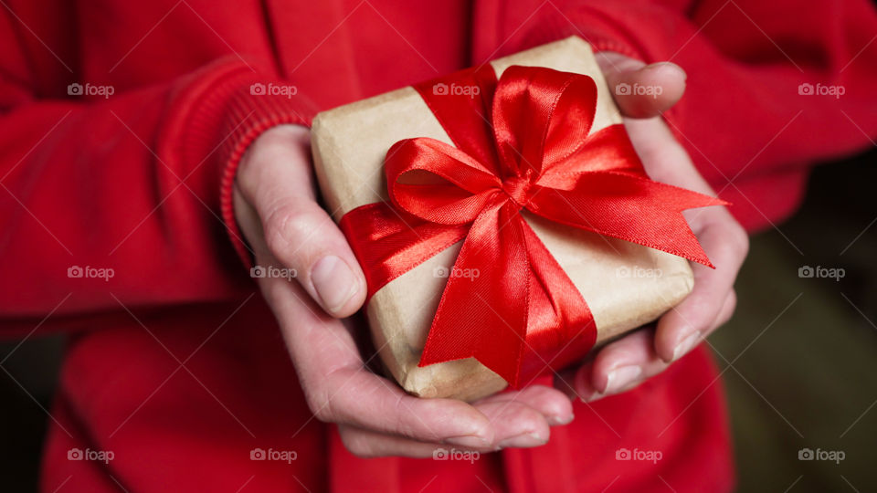 Young woman in red swether holding a gift box wrapped in brown paper and tied with a red ribbon with a bow for a space event. Christmas, New Year, Birthday, Valentine's Day.Close-up.