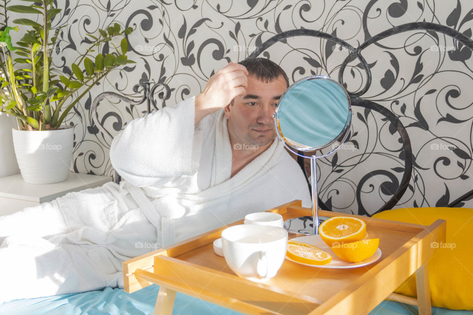 A man and home spa care at home takes care of himself and smears his face with cream, does a manicure at home on a blue background and with oranges and rests.