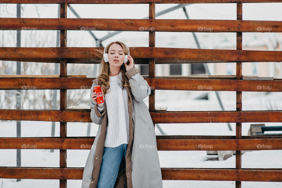 A woman in white headphones listening to music and drinking Coca Cola. Winter cold season. Grey coat. Stylish winter outfit