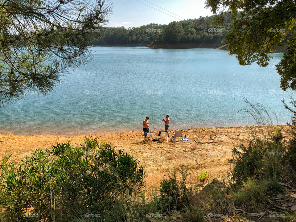 2 guys, viewed from afar, are relaxing at a river beach in Central Portugal. The water looks warm and inviting and they are surrounded by lush foliage 