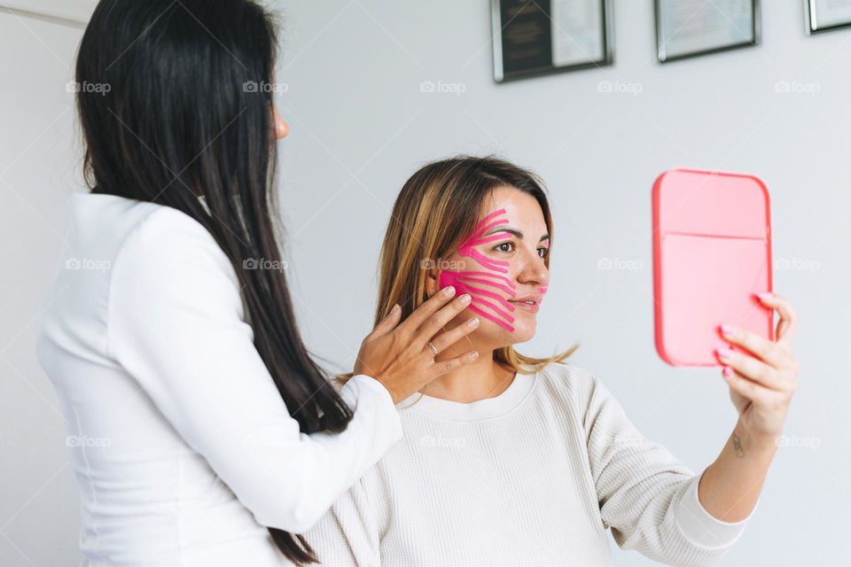 Young beautiful brunette woman with long hair doctor cosmetologist makes face taping for patient in office. Young women do anti-aging procedures with help of kinesio tape