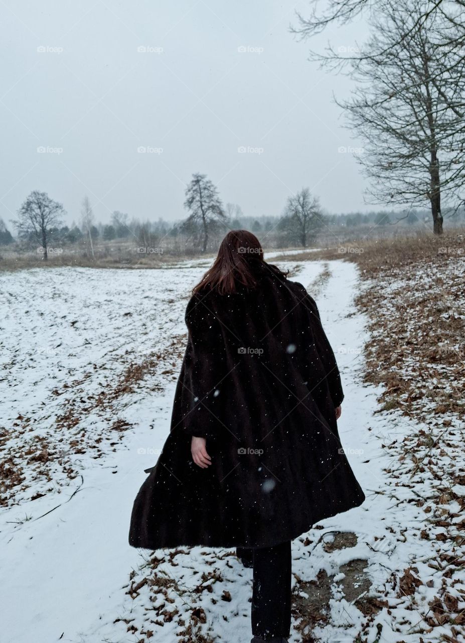 beautiful photo from the back of a girl in a black faux fur coat walking in a snowy field