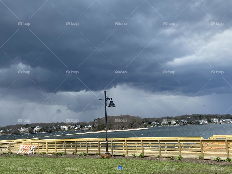 Black clouds above the water on an April afternoon clearly signal a monster storm is coming. The cloud behind the lamppost looks like a dinosaur head with its mouth open, and mostly everyone has heeded the warning and left the area.