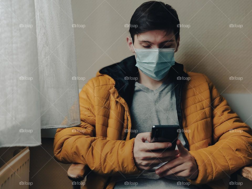 A handsome young Caucasian man in a yellow jacket with brown short hair and a medical mask on his face sits on a chair by the window in the waiting room of a medical office with a black phone in his hands and looks at the screen, reading the news