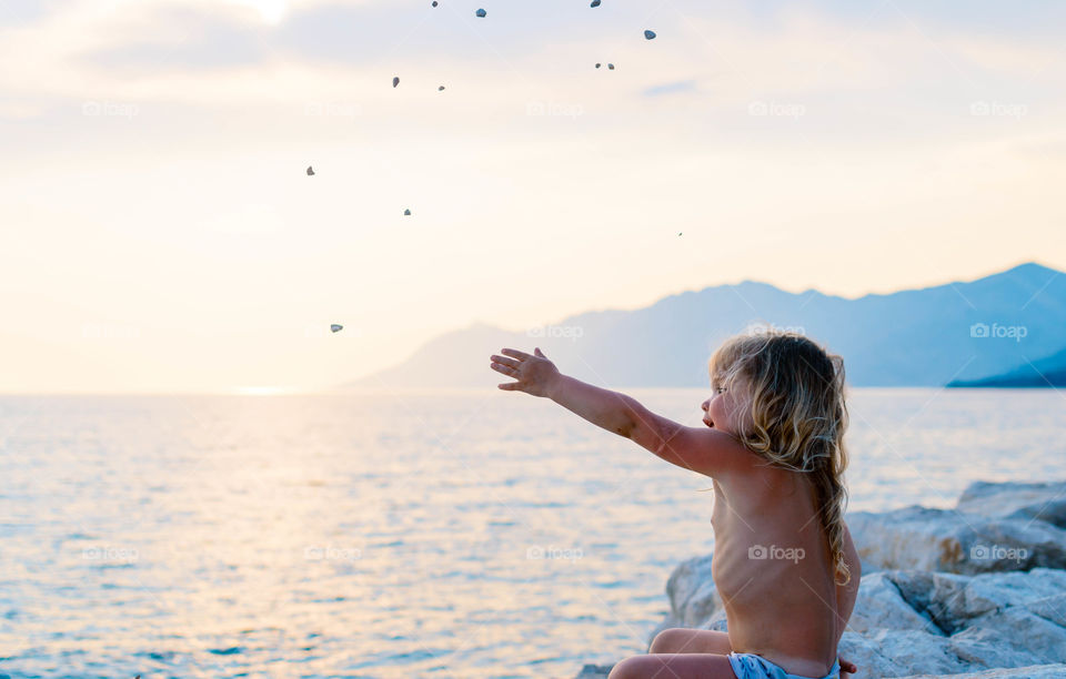 little girl sitting on the sunset beach and throwing pebbles