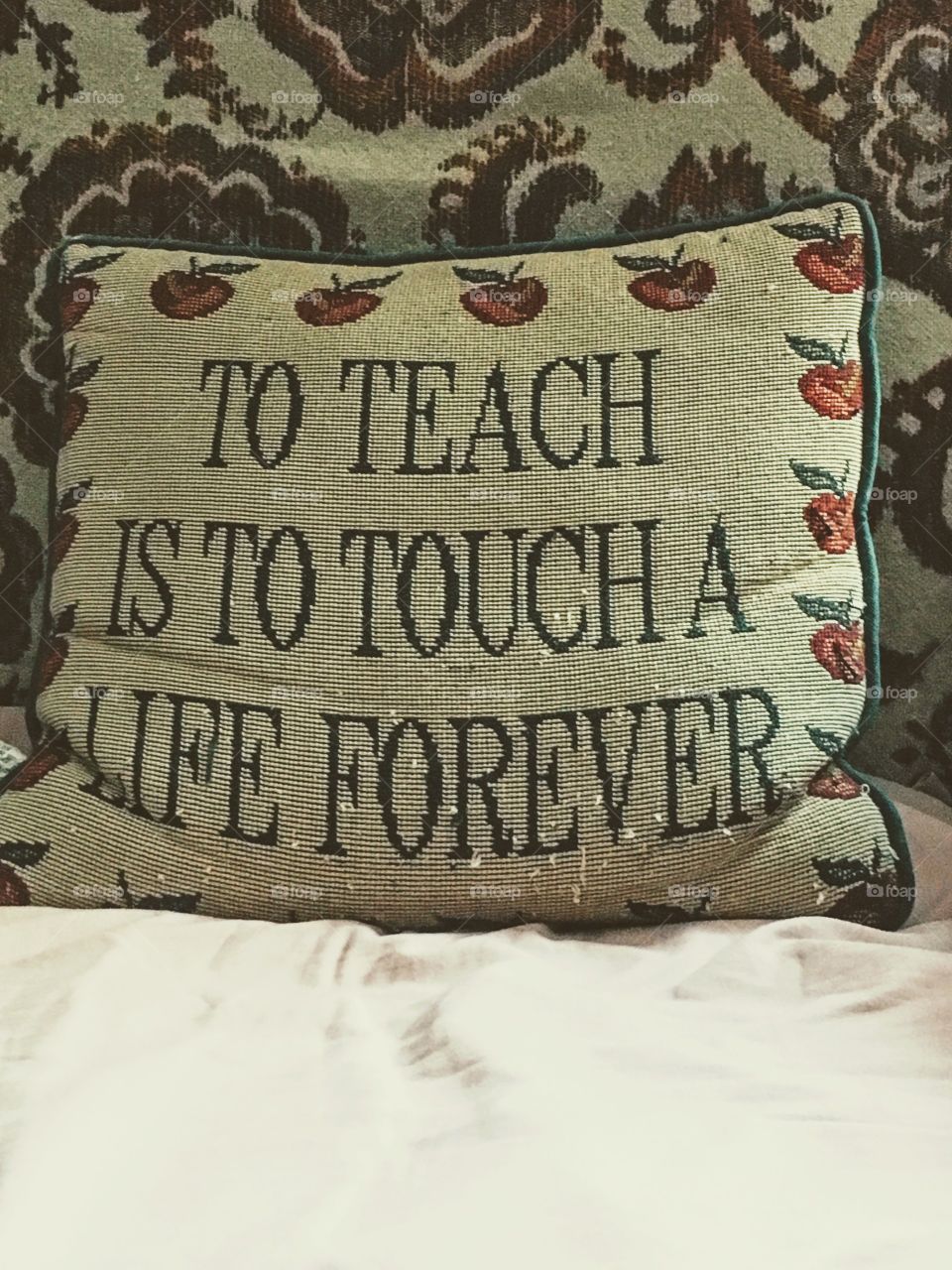 "Teacher's Creed" - Inspiring quote about teachers is printed on a pillow adorned with apples embroidered around the words. Pillow is positioned on a sheet covering a couch with similar color pattern