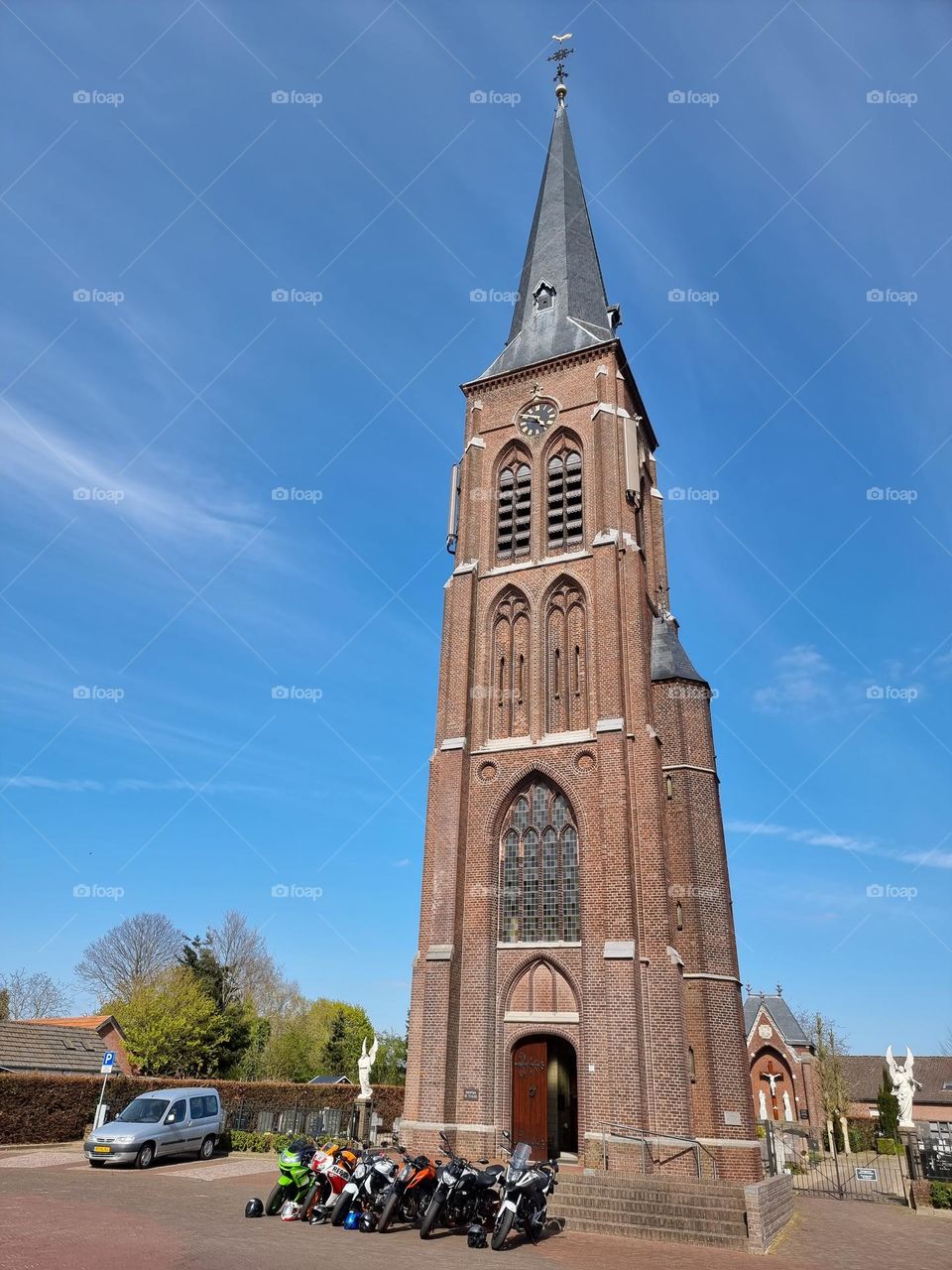 Row of motorbikes parked on the central village square in front of the bell tower of a gothic-style, catholic church on a sunny day