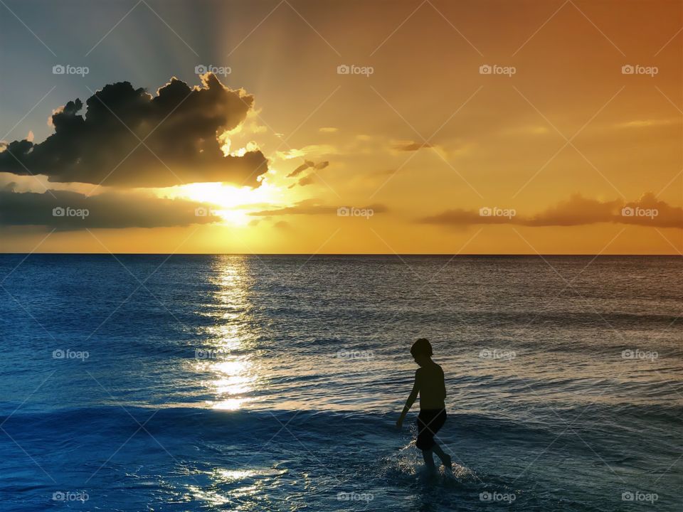 Small boy playing in the ocean waves under a spectacular golden sunset .