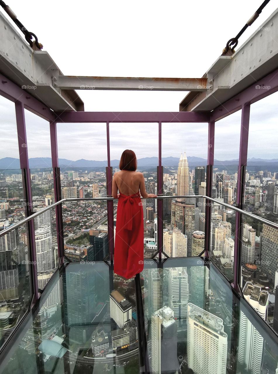 Kuala Lumpur, stunning views of the city and skyscrapers from a height of 300 meters. Observation deck with glass floor. Girl in a red dress.