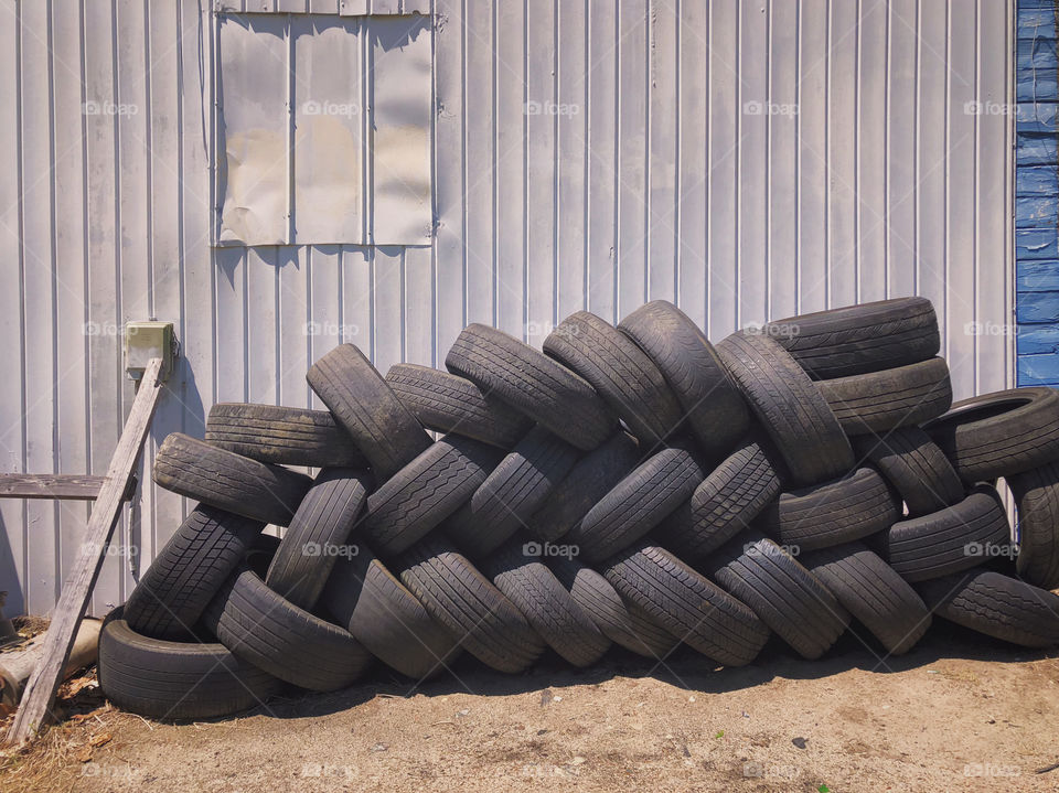 Rustic, in color photo of used tires stacked against an old white wall and in the dirt outside.