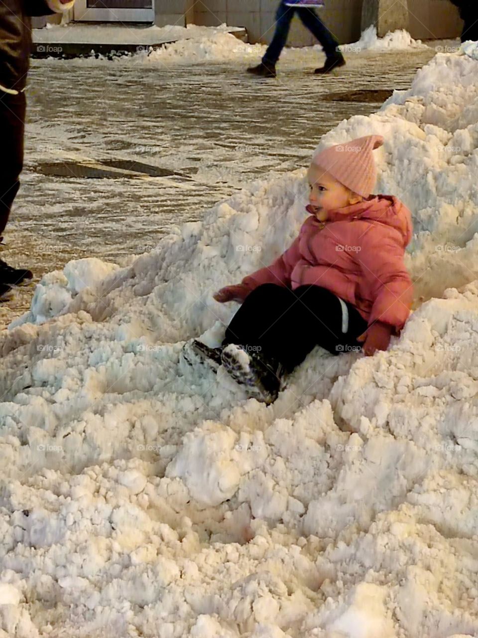 Winter.  There is a small snow slide on the street.  A little girl is riding a snow slide.  Joy and happiness on the girl's face