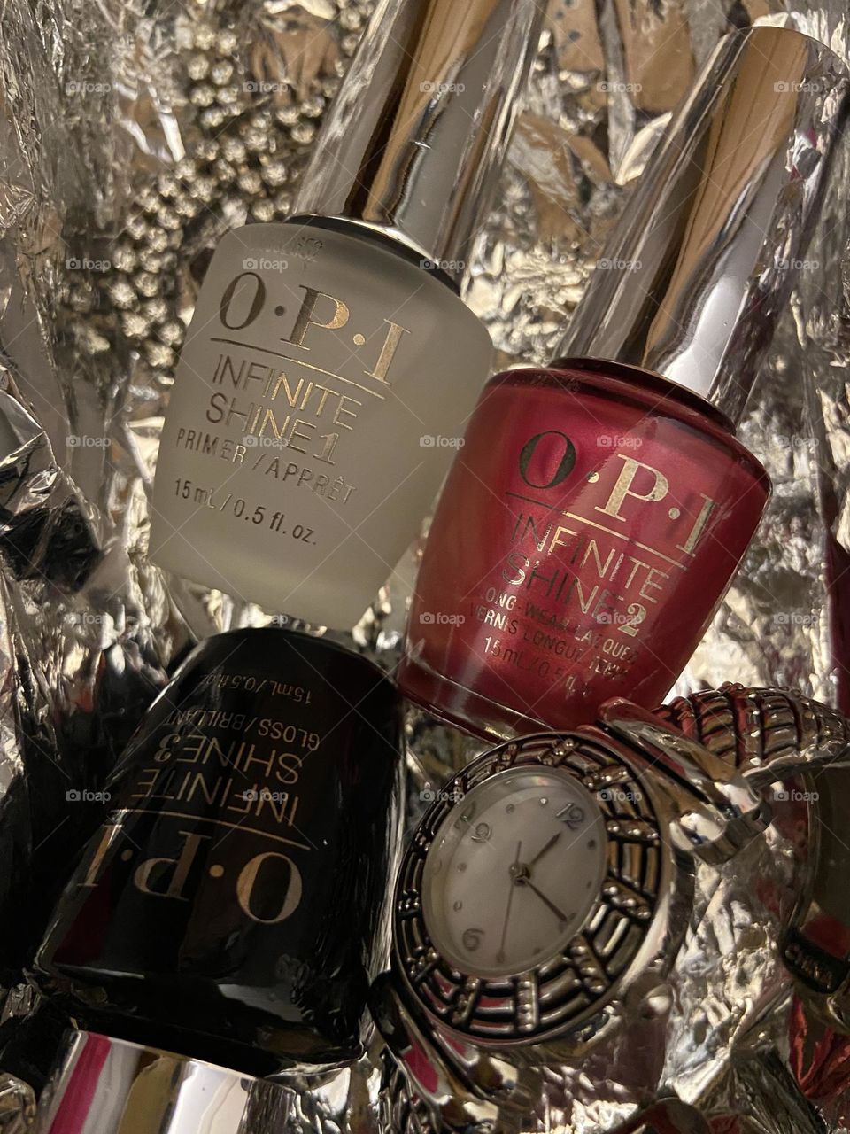 Bottles of Opi Infinite Shine Nail Lacquer displayed with a silver watch set at 12:15 and a silver bracelet twisted into the infinity symbol shape. All on a silver background. The nail polish shade is “15 minutes of flame.” 
