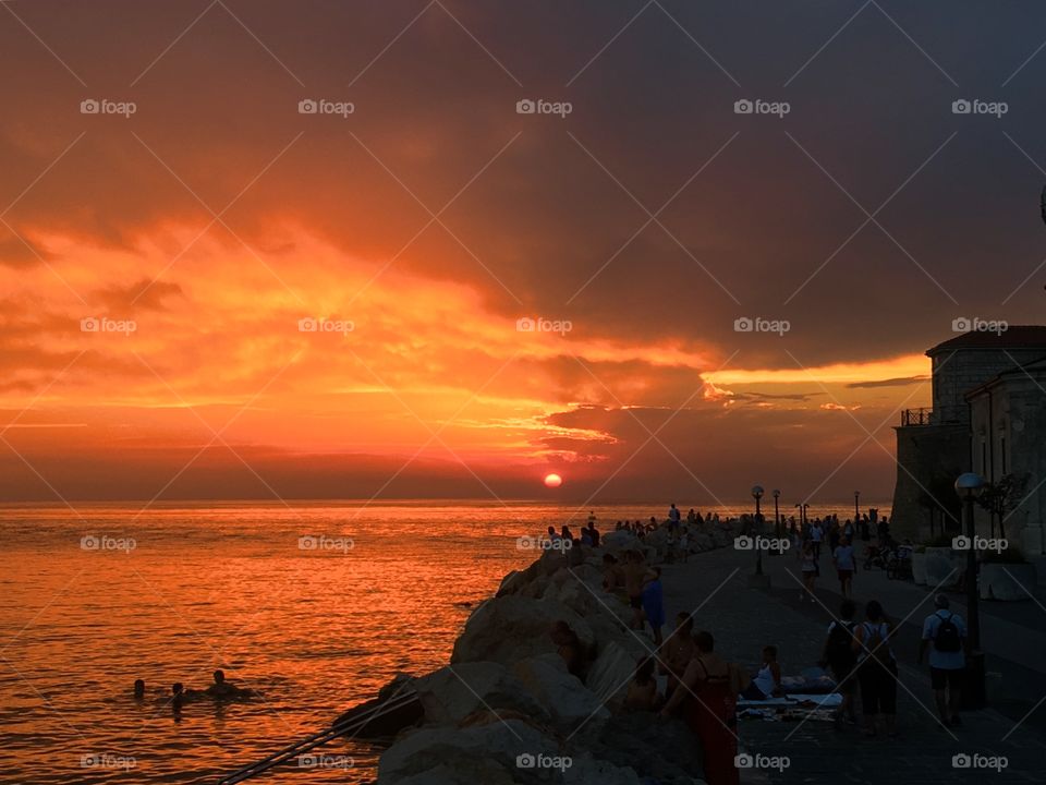 Taken on the coast of the small Slovenian town of Piran, on a summer evening. People can be seen admiring the sunset along the promenade, and also swimming in the sea by the rocky coast. With the intense orange glow of the sun lighting up the scene.