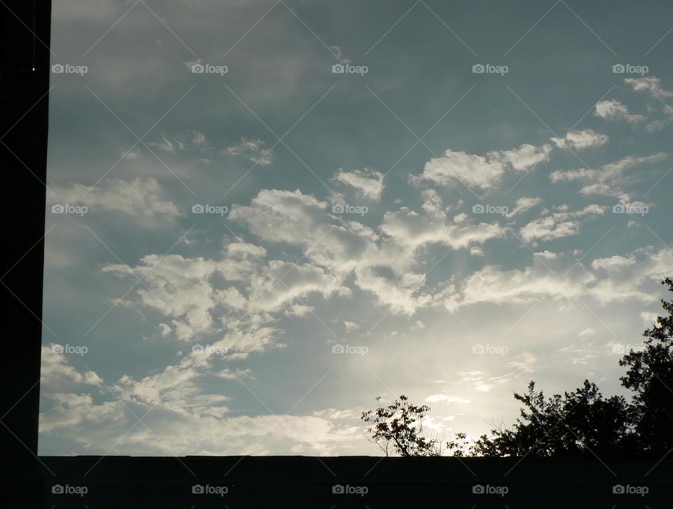 Sunset in the evening creating a shaded area of the house roof and also showing the beauty of the cloudy illuminated sky from the sun and seeing some branches and leaves from trees peaking out above the roof.