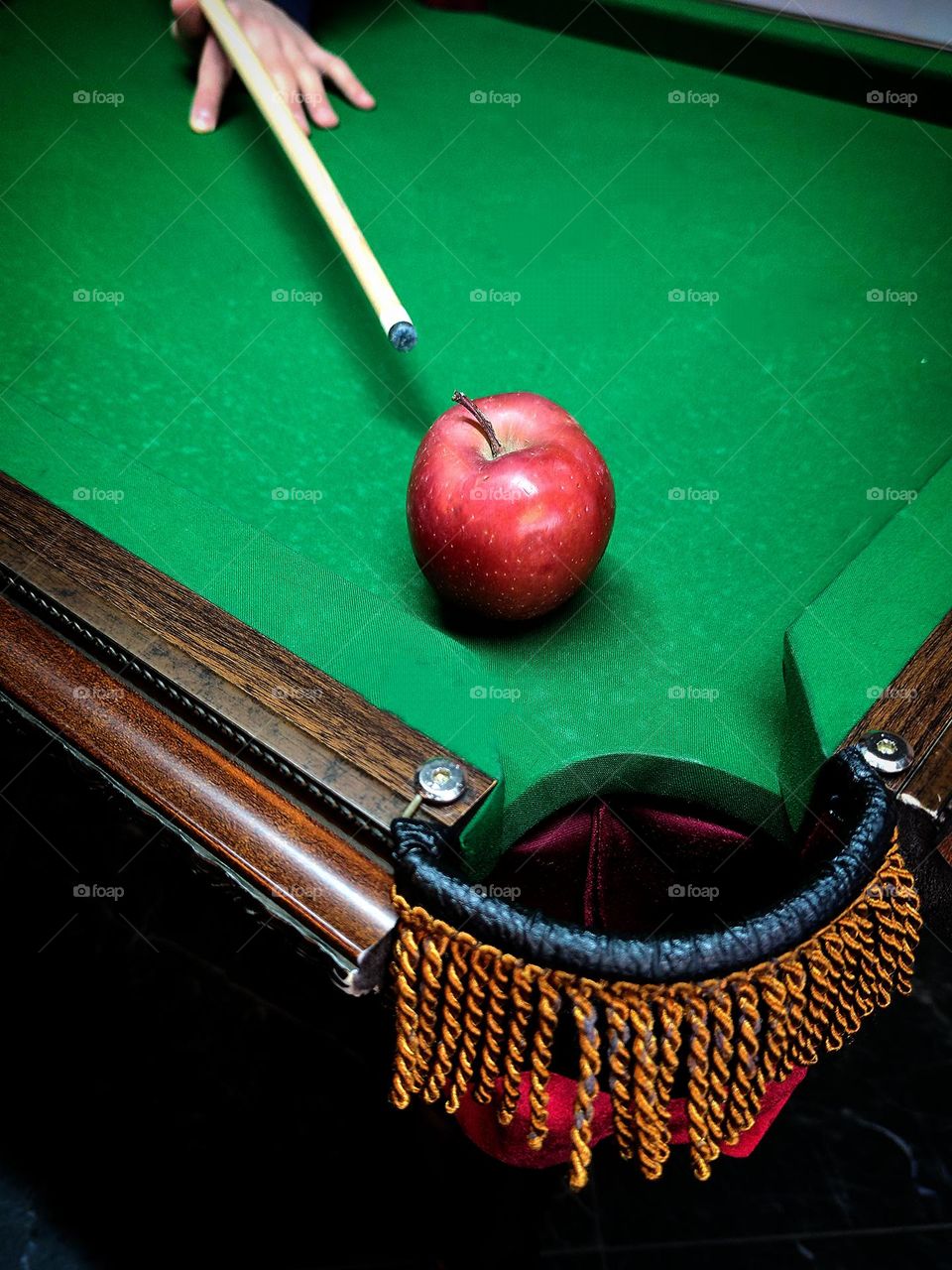 There is a red apple near the pocket on the green billiard table.  In the background is a wooden cue held by a hand to strike.