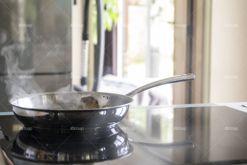 A portrait of a metal frying pan, frying some bacon on an induction furnace. the reflection of the pan is in the glass of the furnace.